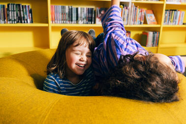 Two children laughing on a sofa, with bookshelves in the background.