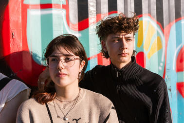 Two young people standing in the sunlight in front of a graffiti wall.