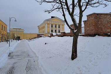 Talvinen maisema, jossa luminen kävelytie kulkee vasemmalla ja oikealla puolella näkyy paljas puu sekä punatiilinen muuri. Taustalla kohoaa vaaleankeltainen, klassistinen rakennus.