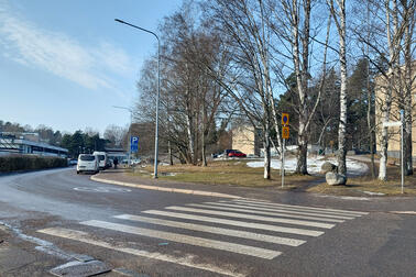 Oikealle kaartuva katu, etualalla on suojatie. Vasemmalla näkyy matala rakennus ja oikealla korkeita koivuja. Taivas on selkeä.