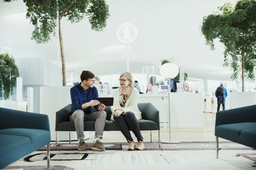 Kaksi henkilö istuu sohvalla kirjastossa ja katselee tablettitietokonetta.