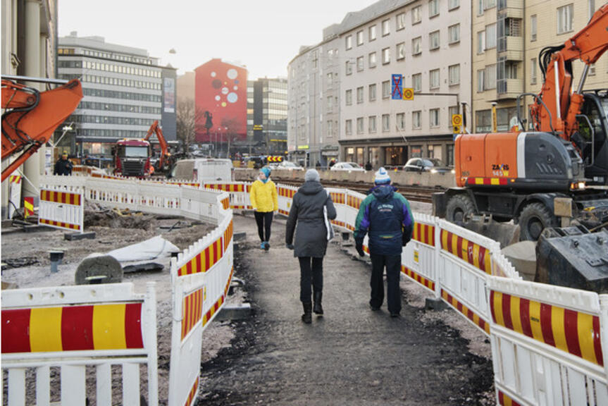 Ihmisiä kävelemässä Kurvin alueella työmaa-aitojen lähellä. Ihmisiä kävelemässä Kurvin alueella työmaa-aitojen lähellä.