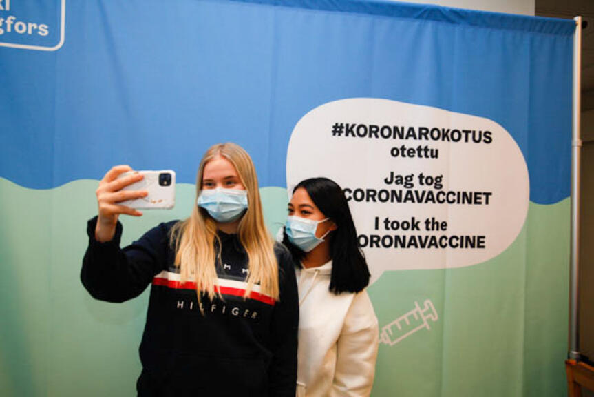 Kaksi tyttöä ottavat kuvan koronarokotus-kankaan edessä. Kaksi tyttöä ottavat kuvan koronarokotus-kankaan edessä.