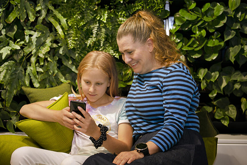 Tyttö lukee mobiilia, äiti katselee hymyillen. Tyttö lukee mobiilia, äiti katselee hymyillen.