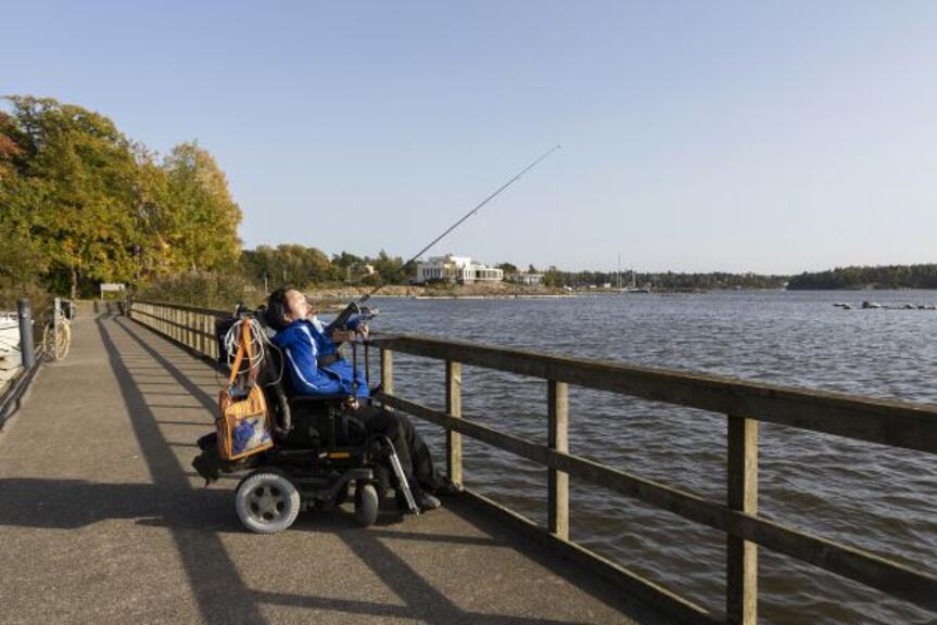 Mies istuu pyörätuolissa ja onkii sillalla. Mies istuu pyörätuolissa ja onkii sillalla.