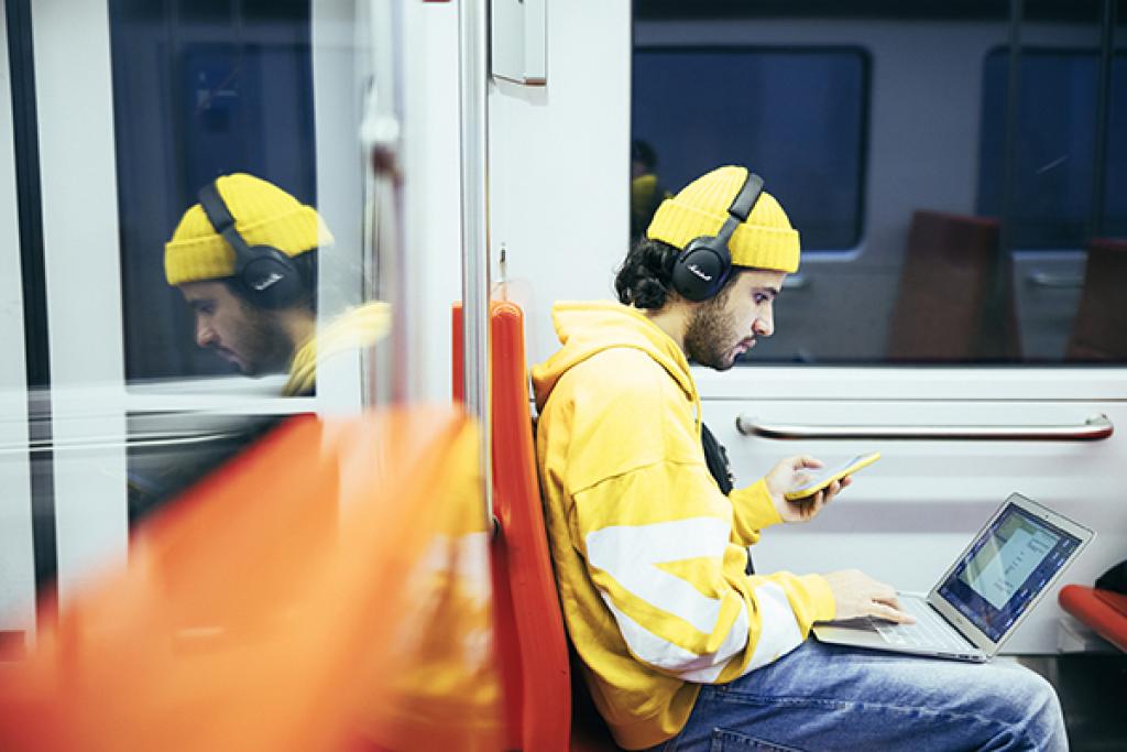 Mies istuu metrossa ja selailee kannettavaa tietokonetta ja puhelinta.