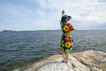 Kuvassa kukkamekkoinen ja lierihattuinen nainen tervehtii käsi ylhäälle merta, seisoessaan kalliolla.