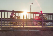 Sillalla aurinkoa vasten pyöräilee kaupunkipyörillä kaksi hahmoa.