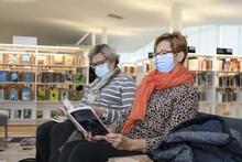 Kaksi henkilöä vierekkäin maskit kasvoilla istumassa kirjastossa.