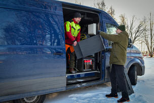 Kaksi miestä nostaa mustaa pönttöä sinisestä pakettiautosta.
