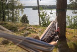 Henkilö makaa riippumatossa. Riippumatto on kiinnitetty puuhun. Taustalla näkyy meri ja purjelaiva. 