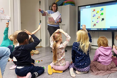 Lapset istuvat lattialla opettajan ja ison näyttöruudun edessä.