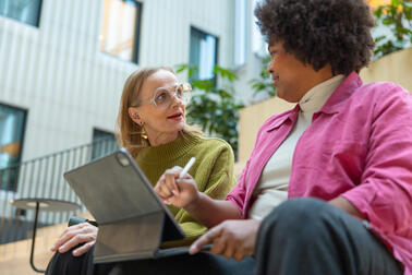 Two women are sitting on stairs, talking. One of them has a tablet in her lap.