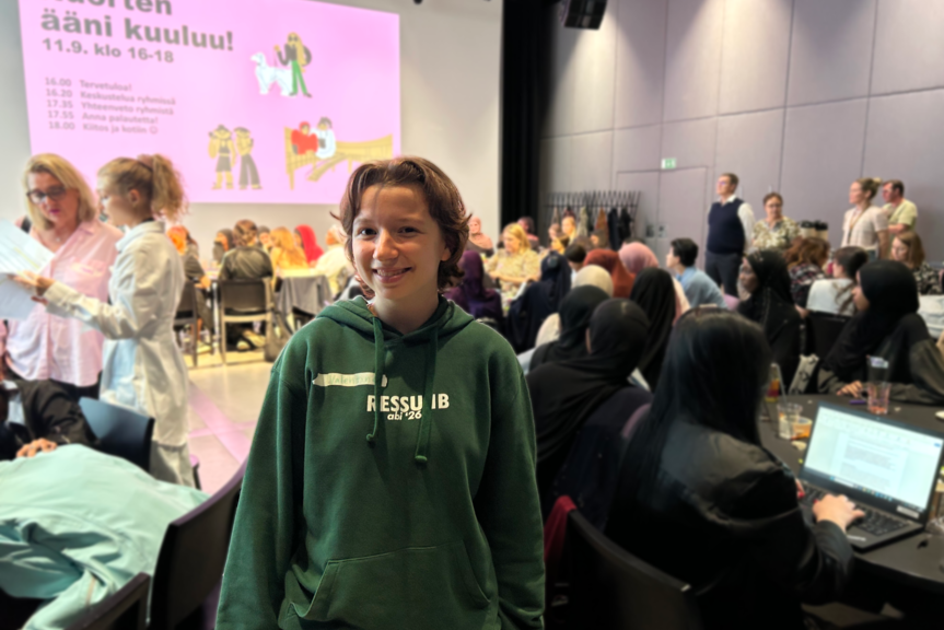 Nuori katsoo kameraan hymyillen isossa salissa, taustalla ihmisiä istuu pöydissä. Takaseinälle on heijastettu esitys, jossa lukee Nuorten ääni kuuluu. 