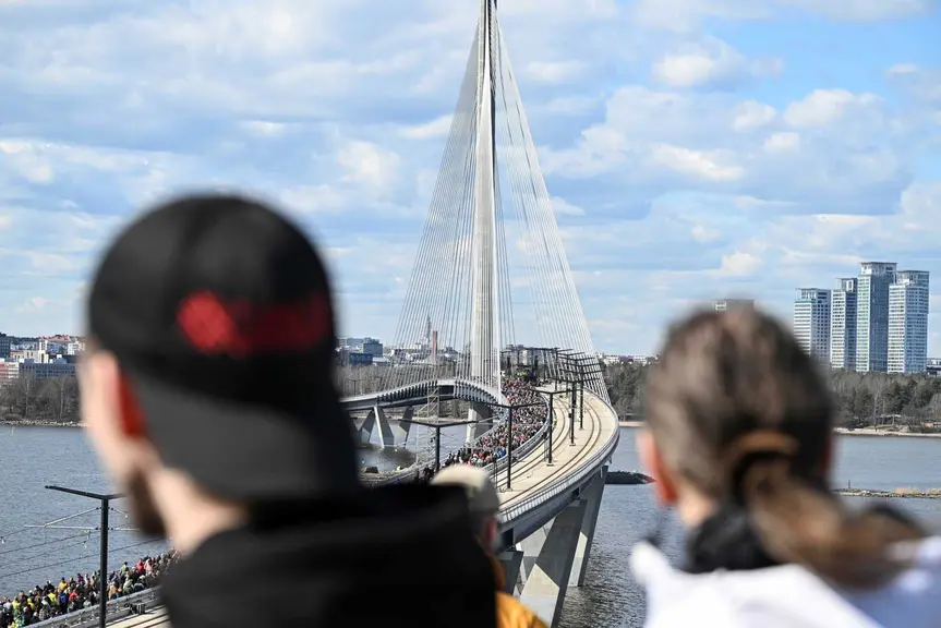 Kruunuvuorensilta kuvattuna etäältä. Sillalla kulkee runsaasti jalankulkijoita, ja etualalla näkyy kaksi epätarkkaa katsojaa. Taustalla näkyy merta ja kaupunkimaisemaa.
