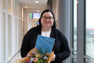 Henkilö seisoo valoisalla käytävällä ja pitää käsissään kukkakimppua sekä sinistä kansiota, jossa on organisaation logo. Taustalla näkyy käytävä ja ikkunoista avautuva näkymä ulos.