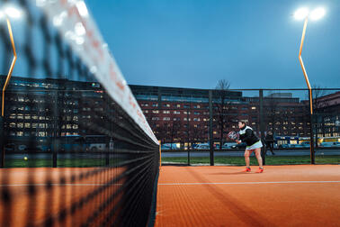 Henkilö valmistautuu ottamaan palloa vastaan kaupunkimiljöössä sijaitsevalla padelkentällä.