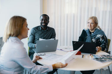 Kolme henkilö istuu tietokoneiden ääressä ja juttelee.