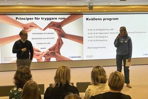 Två personer står framför en presentation och talar till en publik.